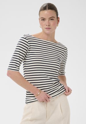 Žena s uhladenými vlasmi, ktorá má na sebe čierno-biely top s lodičkovým výstrihom s pruhmi a béžové nohavice, doplnená zlatými náušnicami vo forme kruhov a náramkom.