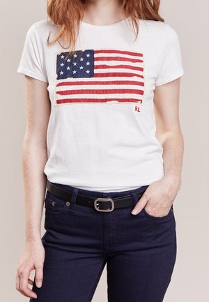 Femme portant un t-shirt blanc avec une grande impression vintage du drapeau américain et un jean bleu foncé avec une ceinture noire, main dans la poche.