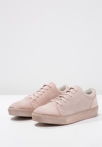 Une paire de baskets en daim rose clair avec des lacets assortis et des semelles en caoutchouc épaisses sur une surface blanche avec un fond uni.