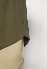 Olive green t-shirt with a straight cut, rounded hem, and a small brand label stitched at the side. Paired with light beige pants.