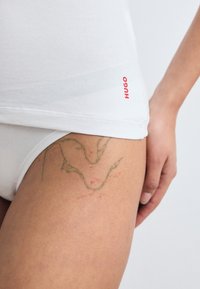Hvite bomullsunderbukser med en rød "HUGO" logo, som passer noe tettsittende. En grønn og rød tatovering er synlig på låret, som avbilder abstrakte former.