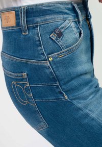 Jeans de denim azul con costuras visibles, una etiqueta de cuero en la parte trasera y un pequeño detalle de bolsillo en el costado. Textura suave y diseño ajustado.