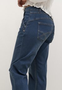 Jeans in denim blu scuro con un design a gamba dritta, tasche posteriori decorate e dettagli di cucitura sottili.