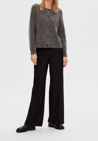 Frau in einer grauen Knopf-Cardigan, schwarzen weiten Hosen und schwarzen Schuhen vor einem einfarbigen hellen Hintergrund.
