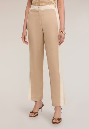 Beige rechte broek met brede witte zijstrepen en gouden knoopdetails, gedragen met bruine sandalen met hak en een gouden armband.