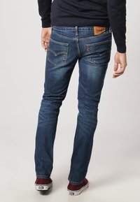 Osoba w ciemnoniebieskich, dopasowanych dżinsach i bordowych sneakersach, stoi plecami do kamery na jednolitym tle.