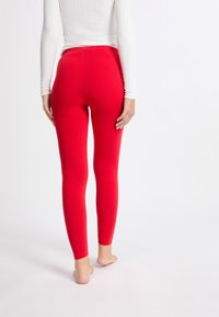 Leggings rojos con textura suave, diseño ajustado y corte a la altura del tobillo. Llevados con una blusa blanca de manga larga; pies descalzos visibles.