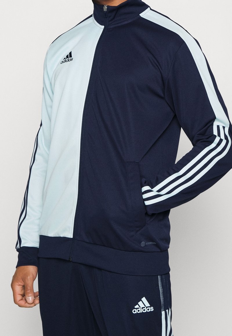 Marinblå och ljusblå zip-up tracksuit-jacka gjord av ett slätt tyg, med tre vita accentlinjer på ärmarna och en dragkedjeficka.
