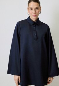 Cămașă oversized de culoare albastru închis, cu guler, plasă cu nasturi și mâneci largi. Fabricată dintr-un material neted cu o textură moale.