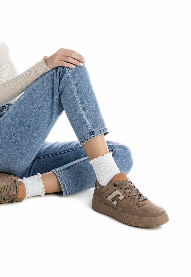 Sneakers in suede marrone con una finitura testurizzata, caratterizzati da un logo prominente, chiusura con lacci e una suola spessa. Indossati con calzini corti bianchi a costine.