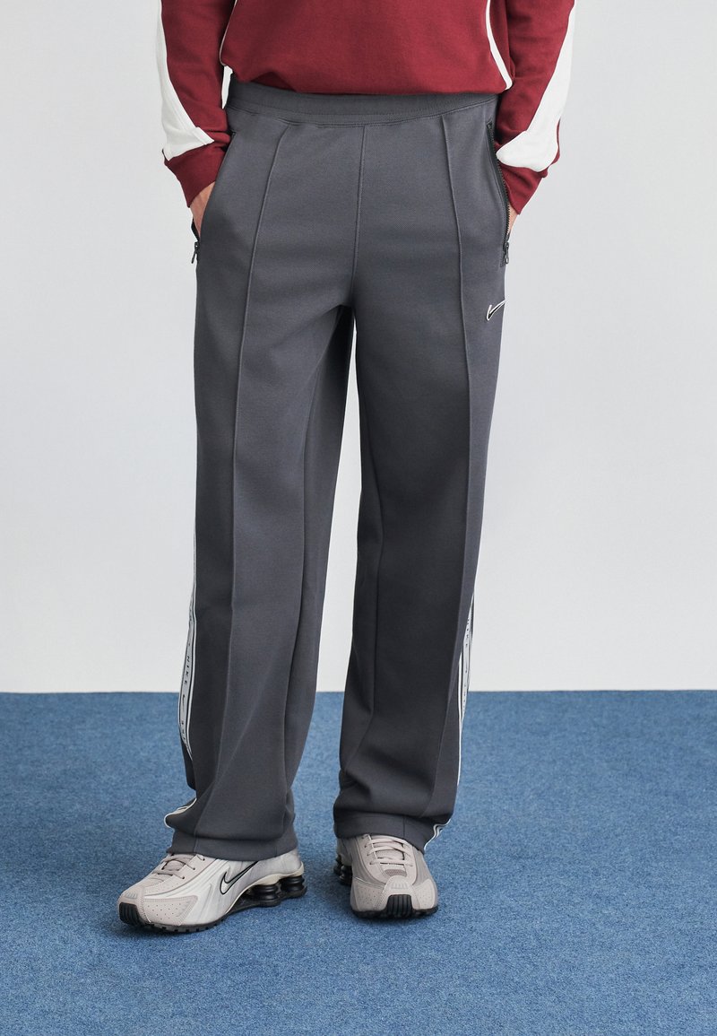 Sive športne hlače z teksturirano površino, z bočnimi zippers in kontrastnimi belimi črtami. Ujemajo se z svetlo sivimi športnimi copati.