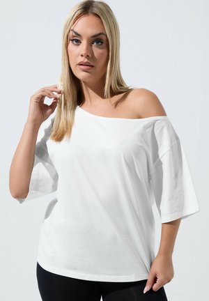 Mujer con cabello largo y rubio que lleva una blusa blanca extragrande sin hombros y pantalones negros sobre un fondo claro y liso.