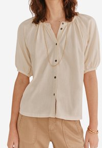 Beige Bluse mit Puffärmeln, V-Ausschnitt, Frontknöpfen und gerafften Schultern. Der Stoff wirkt weich mit einer feinen Textur.