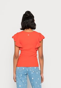 Punainen ribattu yläosa, jossa on röyhelöhihoja, tiukka vartalo-osa ja pyöreä kaula-aukko, yhdistettynä vaaleansinisiin farkkuihin, joissa on päivänkakkukuvioita.