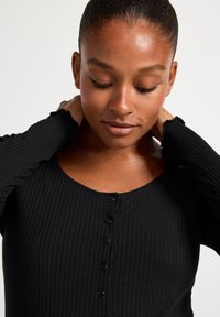 Sort ribbet langærmet top med rund halsudskæring, der har fem knapper foran og flæser ved håndleddene, lavet af et blødt, strækbart materiale.