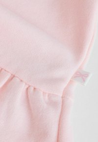 Világos rózsaszínű, puha tapintású anyag, amelyen húzott varrás és egy kis fehér címke található, rózsaszín masni mintával.