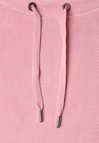 Tissu de sweat à capuche rose avec deux cordons plats roses passés dans des œillets métalliques, chacun se terminant par un embout argenté.