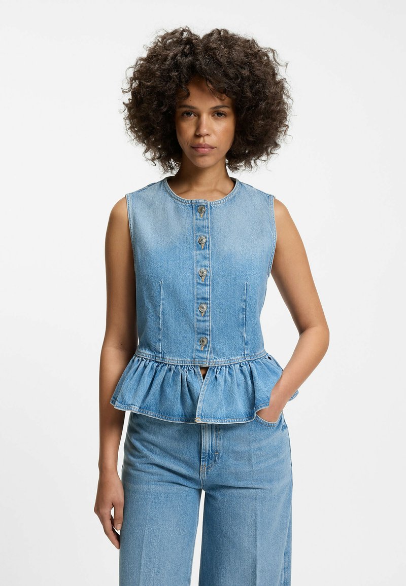 Femme aux cheveux bouclés portant un top en denim sans manches boutonné avec un ourlet volanté et un pantalon en denim taille haute, posant avec une main dans la poche.