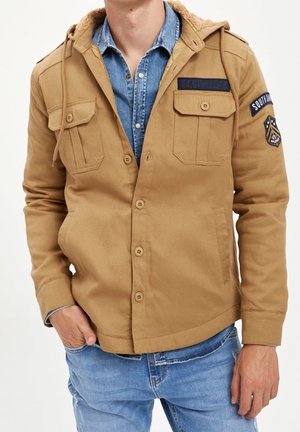 Homme porte une veste marron clair à boutons avec poches poitrine et écussons sur une chemise en jean bleue et un jean, main dans la poche sur fond blanc.