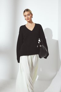 Schwarzer übergroßer Pullover mit V-Ausschnitt, kombiniert mit weißen fließenden Weitbeinen. Hält eine schwarze Handtasche; die glatte Textur steht im Kontrast zu dem Strick.