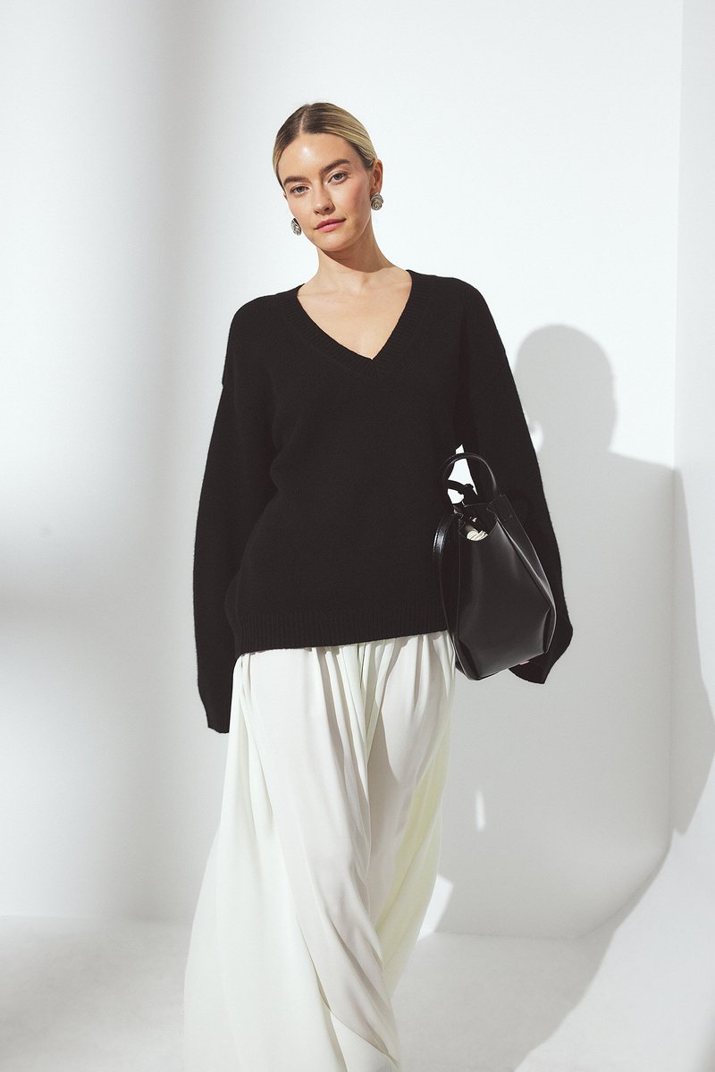 Schwarzer übergroßer Pullover mit V-Ausschnitt, kombiniert mit weißen fließenden Weitbeinen. Hält eine schwarze Handtasche; die glatte Textur steht im Kontrast zu dem Strick.