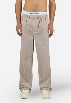 Beige relaxed-fit pants with an elastic waistband featuring a black "PEGADOR" logo. Smooth, lightweight fabric with side pockets and a straight leg.