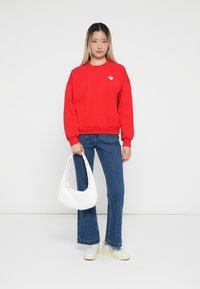 Sweatshirt rouge avec des poignets côtelés, jeans flair bleu clair, sac blanc avec des accents studded, et baskets blanches, se tenant devant un fond blanc.
