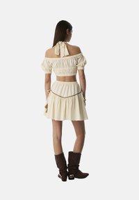 Crop top cremoso con spalle scoperte e maniche a sbuffo, vita arricciata, abbinato a una gonna a campana coordinata. Stivali marroni sopra il ginocchio completano l'outfit.