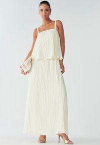 Una donna sta in piedi in un vestito leggero e fluido di colore chiaro con spalline sottili, tiene in mano una piccola borsa e indossa eleganti tacchi dorati.