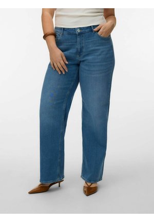 Donna che indossa jeans blu a vita alta a gamba dritta, top bianco, bracciale marrone e scarpe con tacco marrone e punta a punta, in piedi con una mano in tasca.