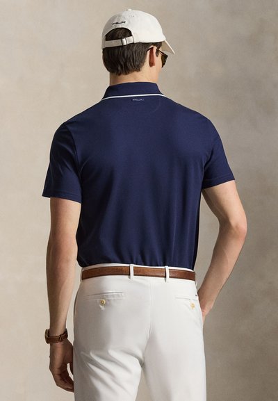 Άνδρας που φορά μπλε σκούρο πόλο, άσπρο παντελόνι με καφέ ζώνη, άσπρο καπέλο και γυαλιά ηλίου, ποζάροντας με την πλάτη σε ουδέτερο φόντο.