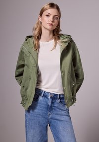 Mladá žena s blond vlasy, která má na sobě stříbrné kruhové náušnice, světle zelenou bundu s kapucí přes bílou košili a džíny s vysokým pasem.