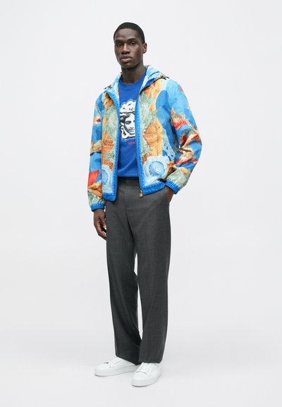 Modra vzorčasta jakna z umetniškimi motivi, nošena čez modro grafično majico in sive hlače, kombinirana z belimi supergami.