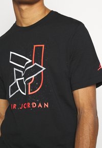 Černé bavlněné tričko s šedým a červeným grafickým designem písmen "J" a "R. JORDAN" na pozadí s efektem rozstřiku.