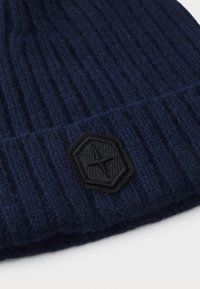 Navy blue knitted beanie with ribbed texture, featuring a black hexagonal logo patch. Material appears soft and warm.