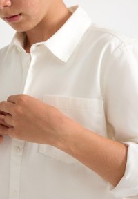 Hvid skjorte med knapper, krave, en enkelt brystlomme og opkrogede ærmer. Glat stoftekstur og skræddersyet pasform.