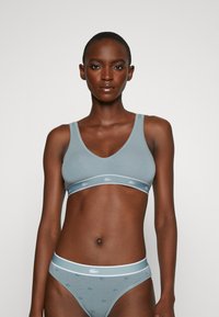 Gaiši zila bralette un bikinī komplekts no mīksta auduma. Bralette ir ar V veida kakla izgriezumu un logo joslu; bikinī ir ar baltu jostu un maziem logo.