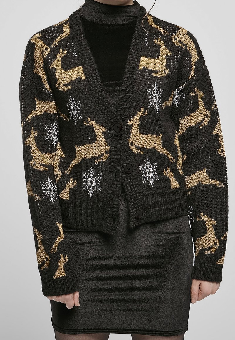 Cardigan en tricot noir avec des motifs de cerfs dorés et des flocons de neige blancs. Il a un col en V, des boutons et des manches texturées.