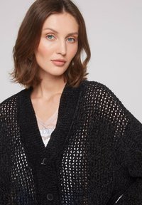 Cardigan noir en tricot avec un design ajouré, présentant un décolleté en V profond et deux boutons à l'avant, superposé sur un haut léger.