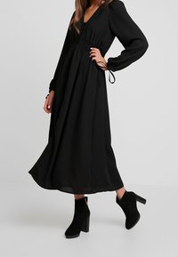 Schwarzes langärmeliges Kleid mit V-Ausschnitt, gerafftem Taillenbereich und fließendem Rock. Kombiniert mit schwarzen Wildleder-Ankelboots mit Blockabsatz.