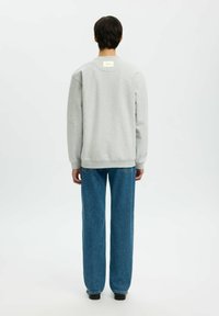 Personne debout de dos, portant un sweat-shirt gris clair et un jean droit bleu, sur un fond blanc uni.