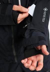 Eine Hand schiebt eine weiße Karte in eine mit Reißverschluss verschlossene Tasche am linken Arm einer schwarzen Gore-Tex-Jacke mit Klettverschlussmanschetten.