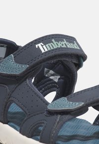Marinblå sandaler med justerbara remmar, med en texturerad ovandel, vadderad sula och en lätt sula. Timberland-logotyp synlig på remmen.