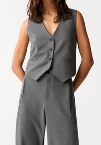 Gilet grigio su misura con scollatura a V, cinque bottoni e senza maniche. Presenta tasche laterali ed è abbinato a pantaloni grigi coordinati.