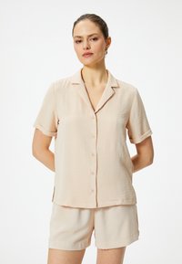 Lichtbeige, kortärmige shirt met een kraag, een borstzak en knoopsluiting, in combinatie met bijpassende beige shorts. Textuurdetail in de stof.
