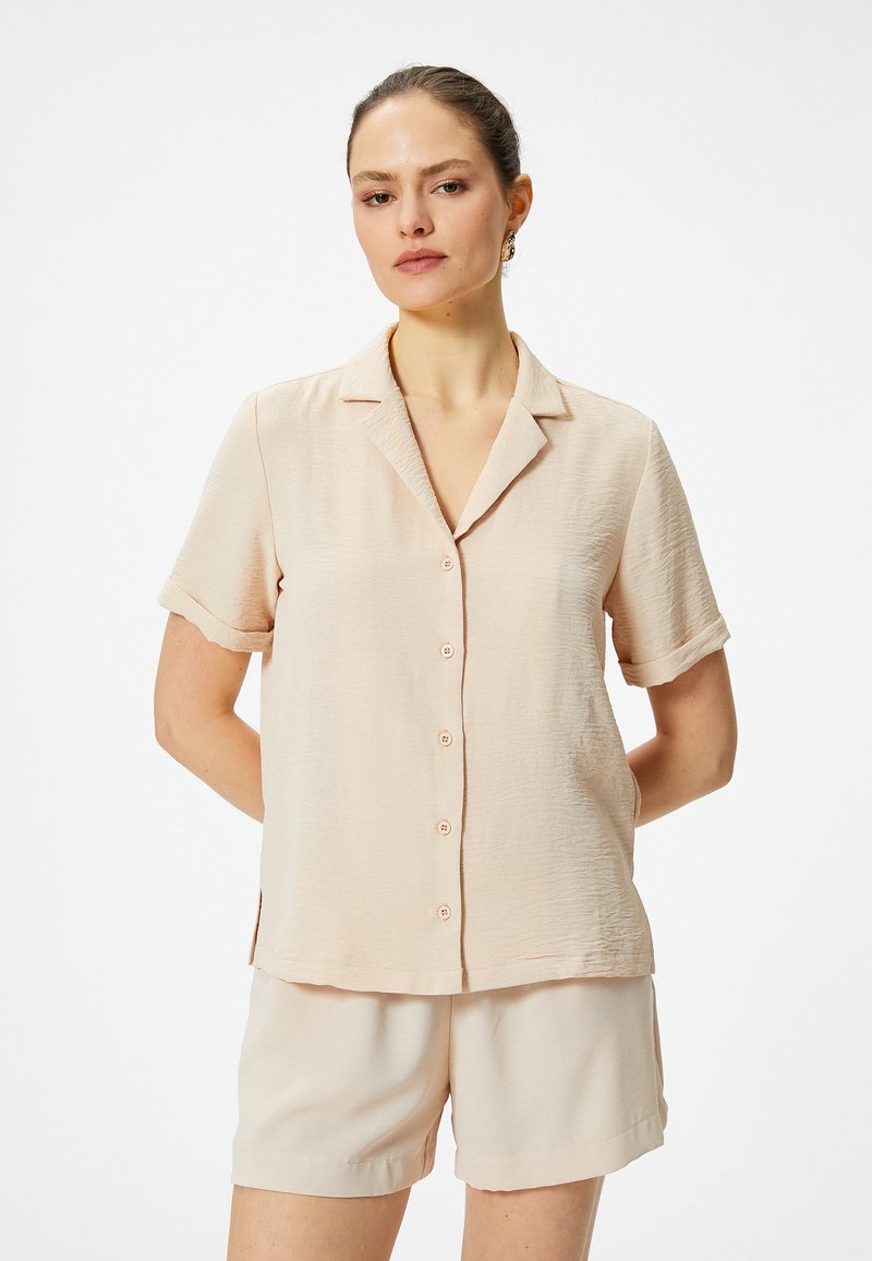 Lichtbeige, kortärmige shirt met een kraag, een borstzak en knoopsluiting, in combinatie met bijpassende beige shorts. Textuurdetail in de stof.