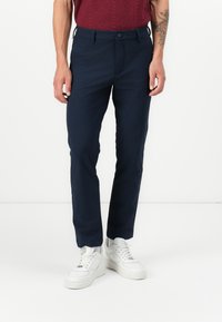 Navy colored trousers with a slim fit, made of smooth fabric, featuring a button closure and side pockets. Worn with white sneakers.