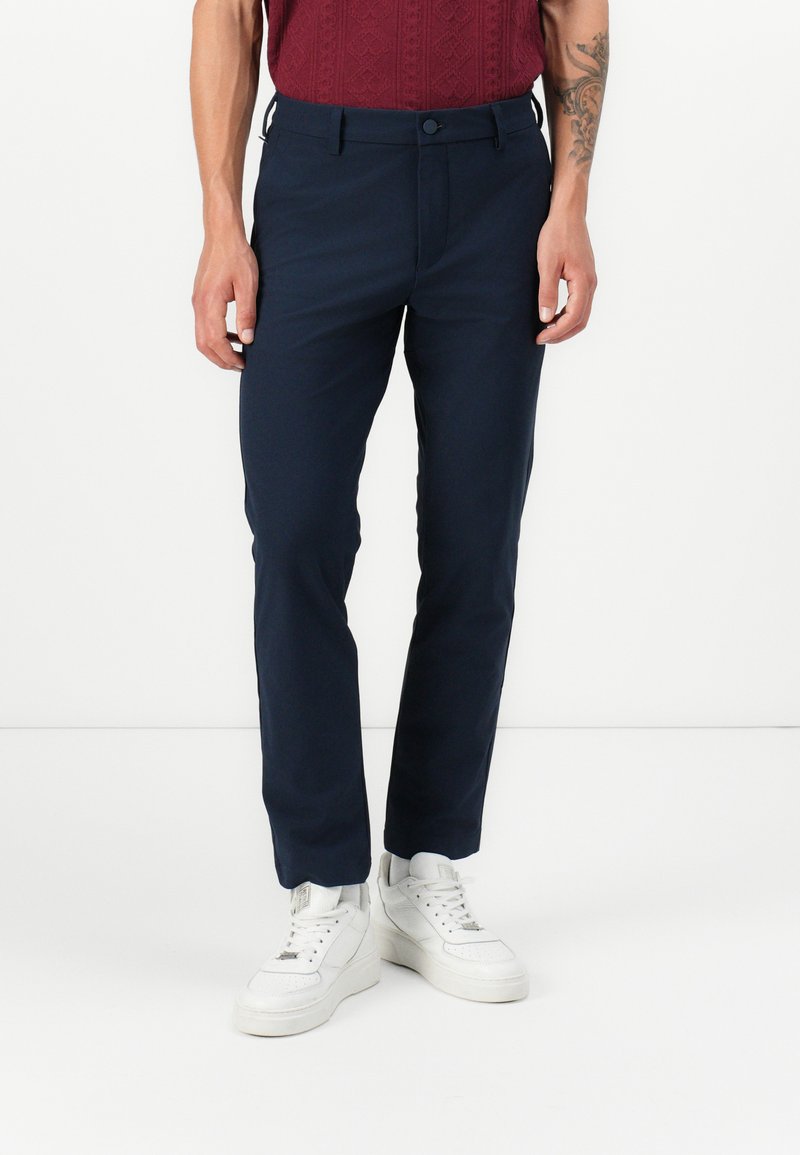 Pantalons de couleur marine avec une coupe slim, en tissu lisse, dotés d'une fermeture à bouton et de poches latérales. Portés avec des baskets blanches.