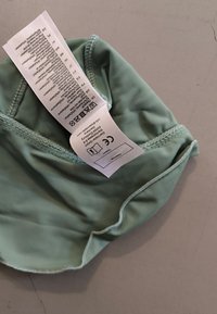 Eine weiche, mintgrüne Stoffmütze mit gerafften elastischen Kanten und einem angehängten Pflegeetikett. Die Textur ist glatt und leicht glänzend.