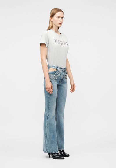 Γυναίκα που στέκεται, φορώντας λευκό πουκάμισο με κοντά μανίκια και μπλε τζιν με διακοσμητικά καρφιά και πλευρικές περικοπές, συνδυασμένα με μαύρα παπούτσια με τακούνι.